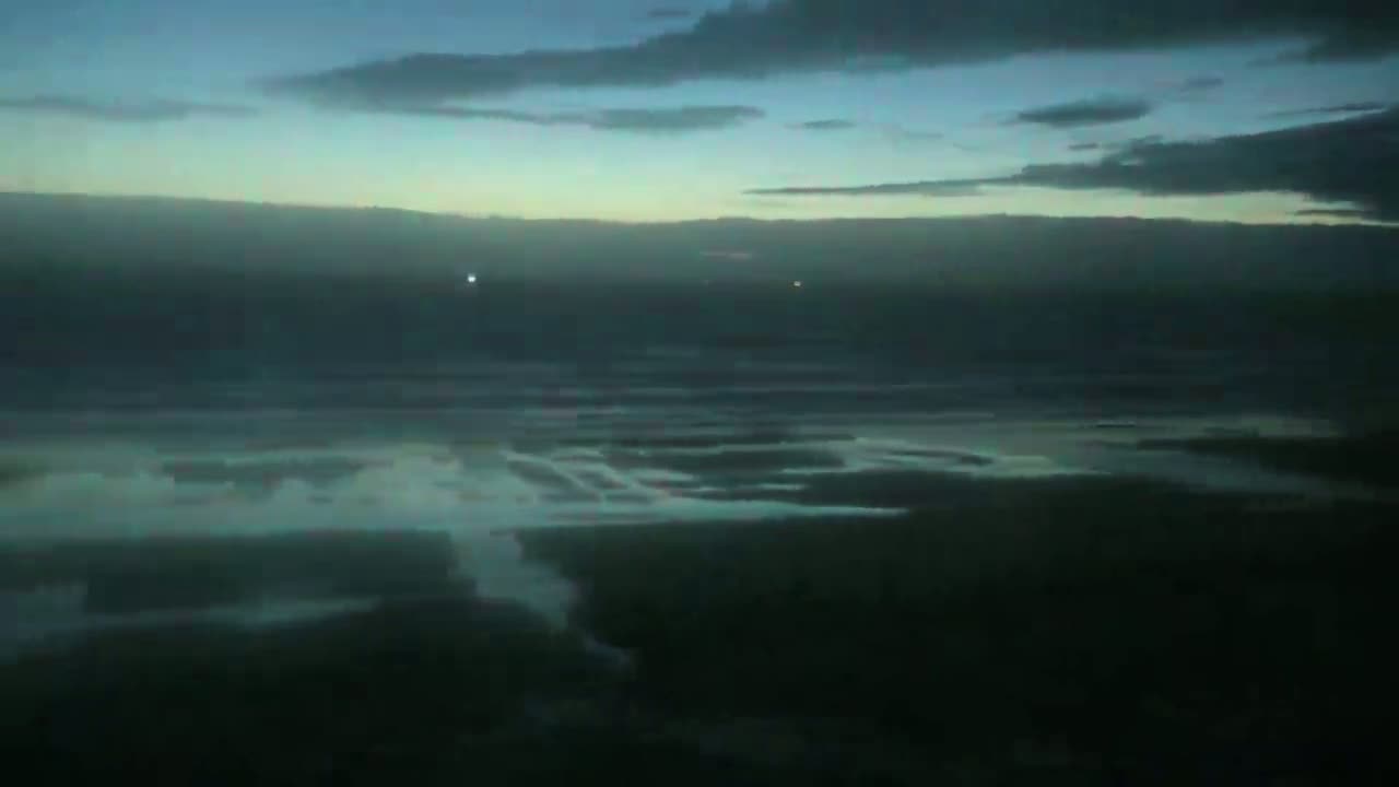 A dark, overcast sky hangs over a flat, dark expanse of water with subtle ripples and a few distant lights.