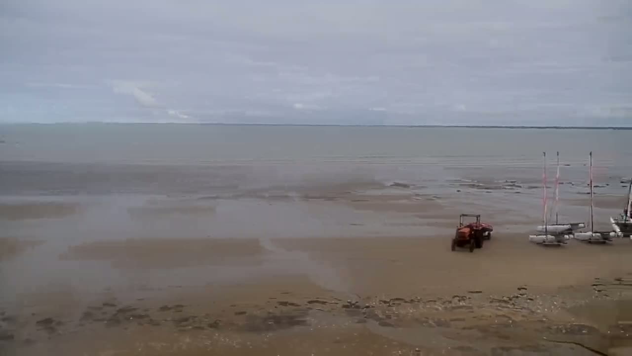 A tractor and several small sailboats are parked on a wide, flat, sandy beach next to calm, grey water under an overcast sky.