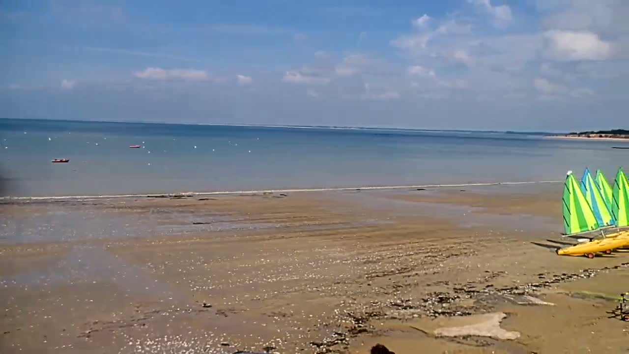 A sandy beach with several small sailboats on the shore and a calm, blue ocean under a partly cloudy sky.