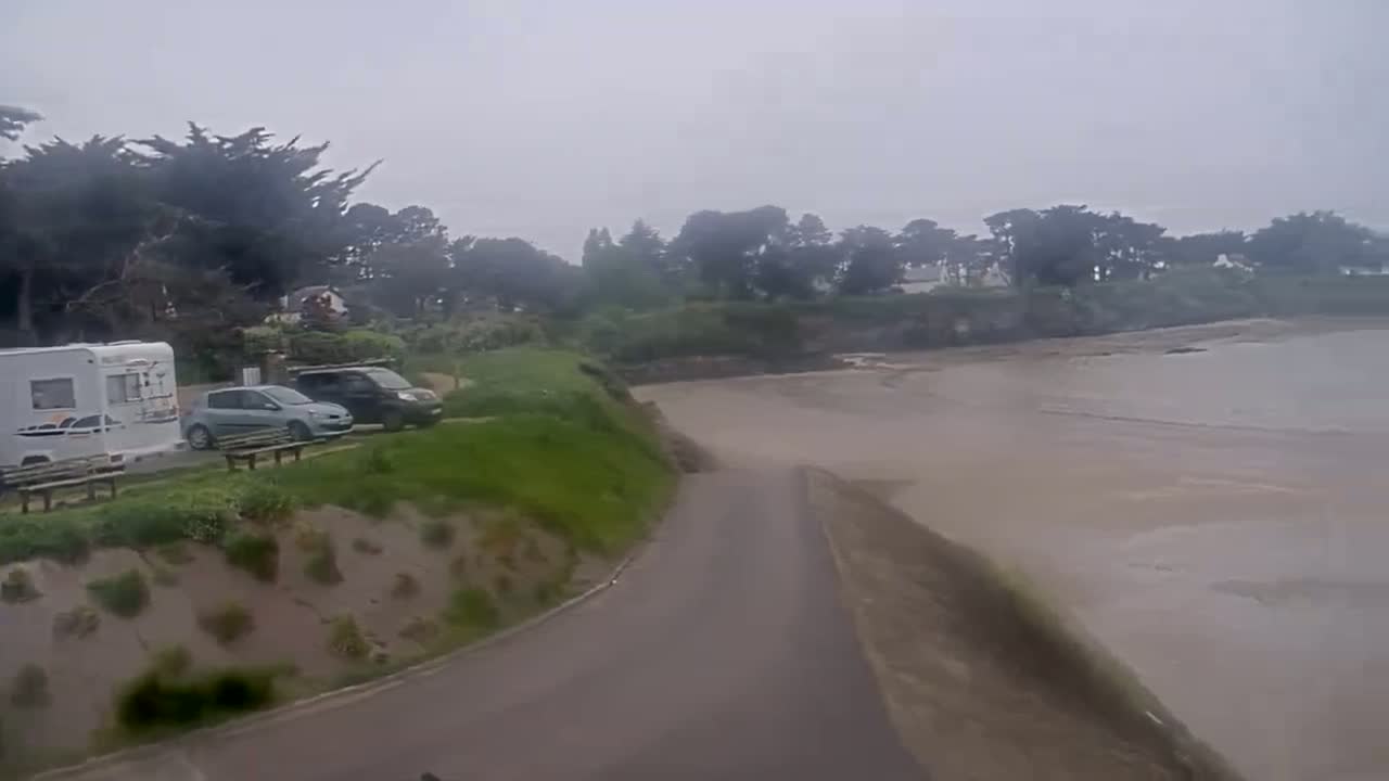 A campervan and several cars are parked near a grassy area overlooking a flat, muddy, and overcast beach.