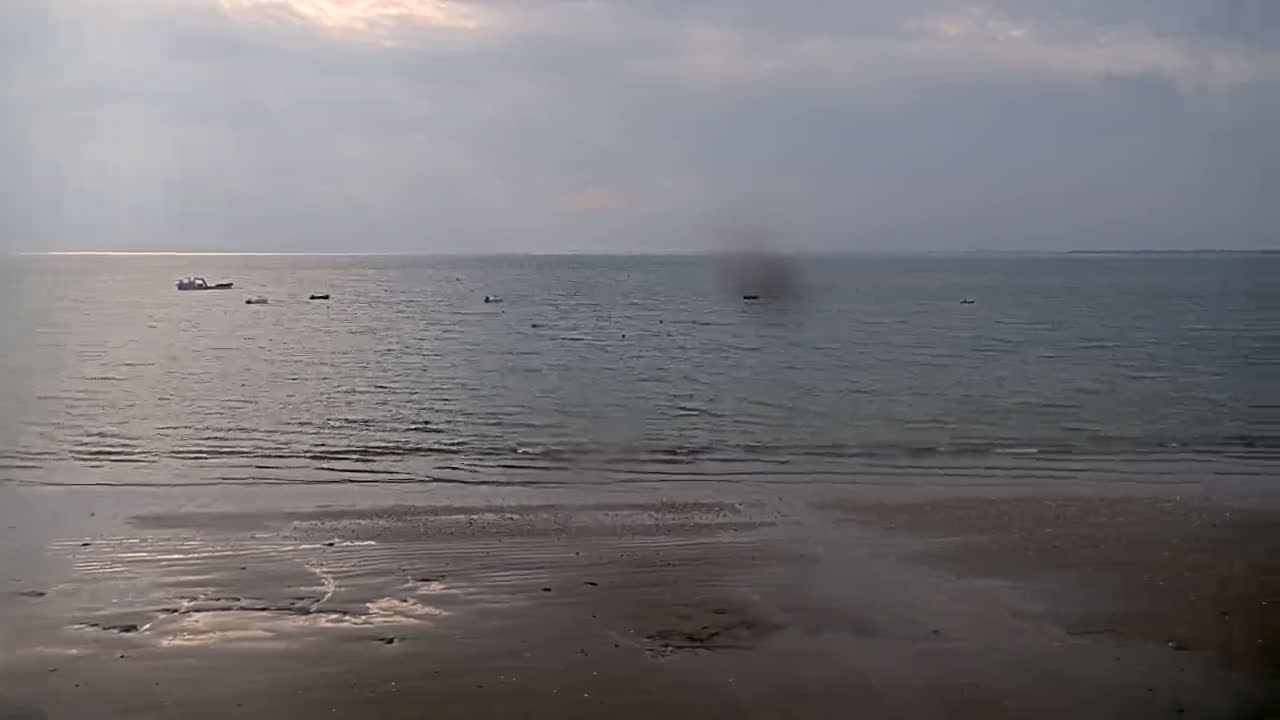 A flat, sandy beach slopes down to a calm sea where several small boats are visible under an overcast sky.