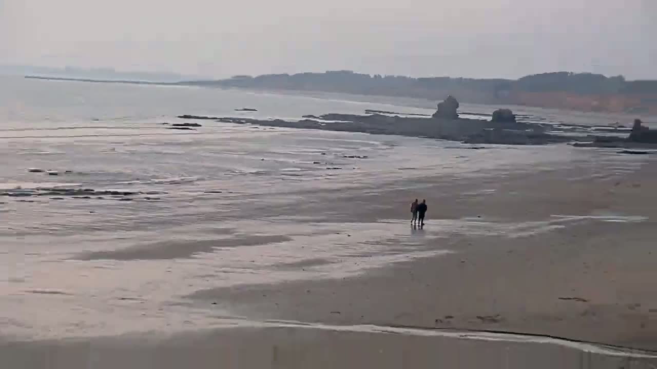 Two people walk on a wide, wet sandy beach near dark rocks and a calm, hazy ocean under an overcast sky.