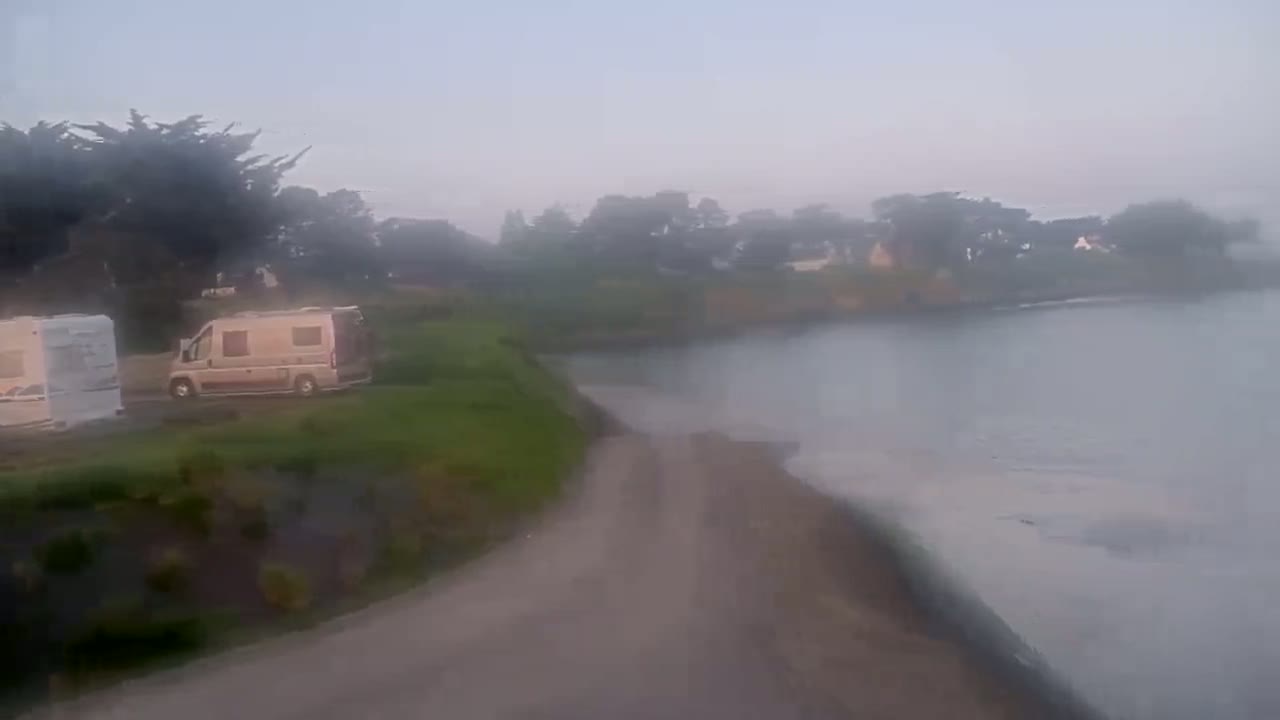Two camper vans are parked near a calm body of water on a foggy morning, next to a grassy area with trees in the background.