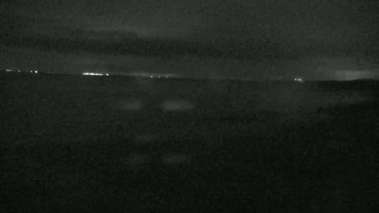 A nighttime, low-light image shows a dark landscape with a distant line of faint lights under a mostly cloudy sky.