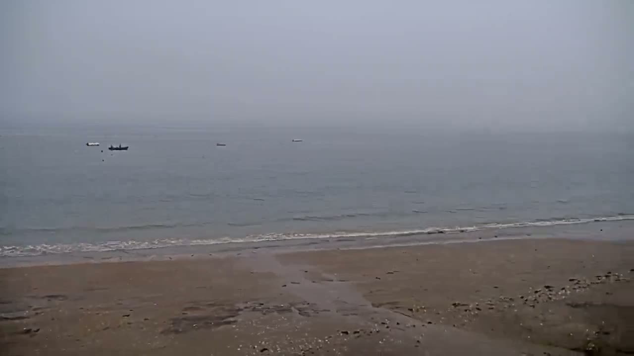 A hazy, overcast day shows a calm sea with several small boats and a wide, sandy beach.