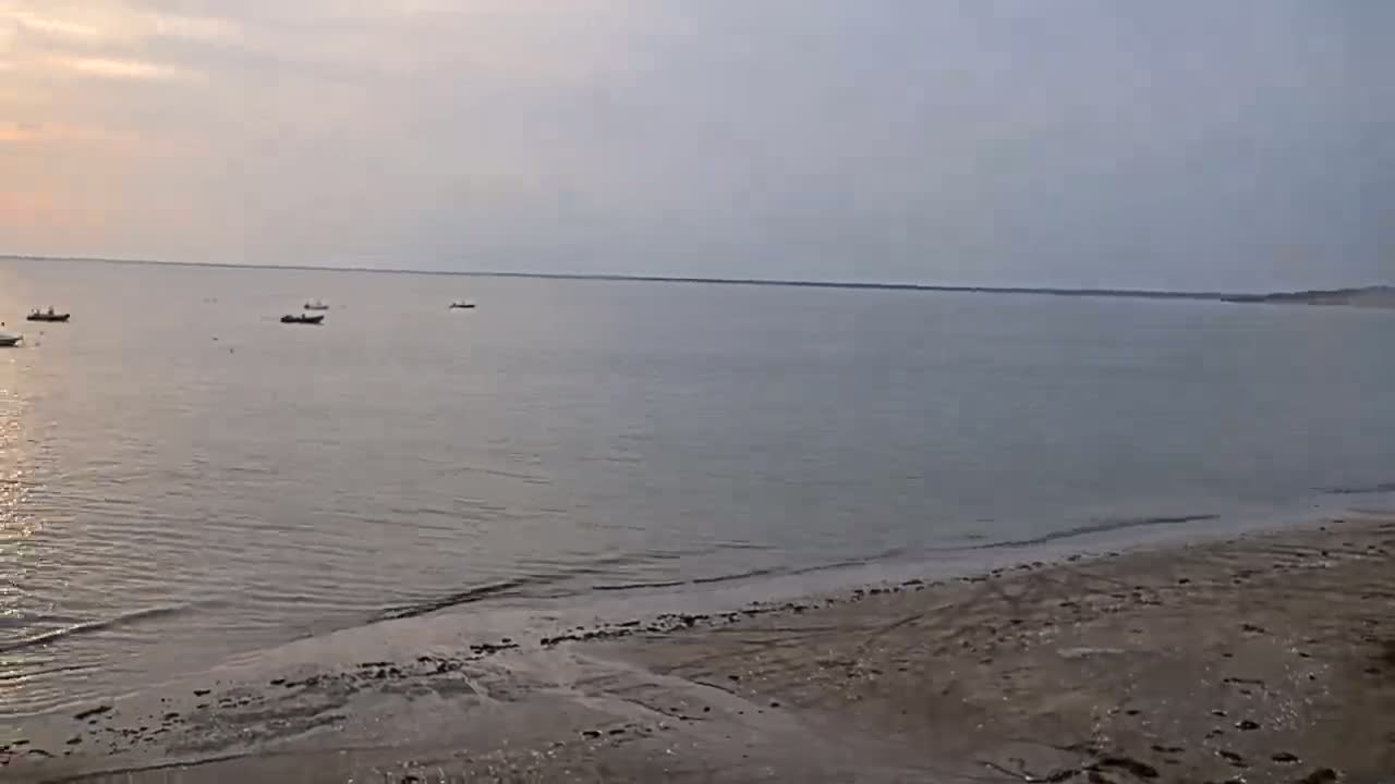 Several small boats are visible on a calm, light-grey sea under a pale, overcast sky, with a sandy beach in the foreground.