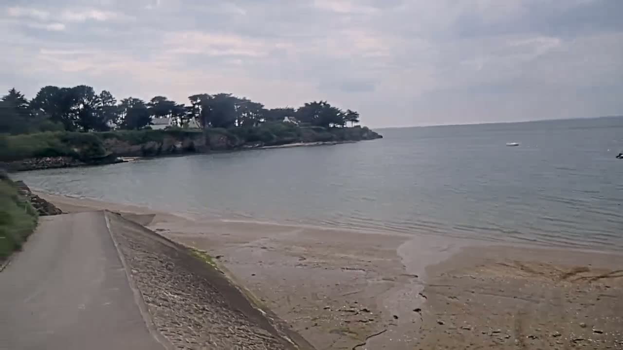 A paved road leads down to a sandy beach and calm bay under an overcast sky, with a small strip of land and trees visible across the water.