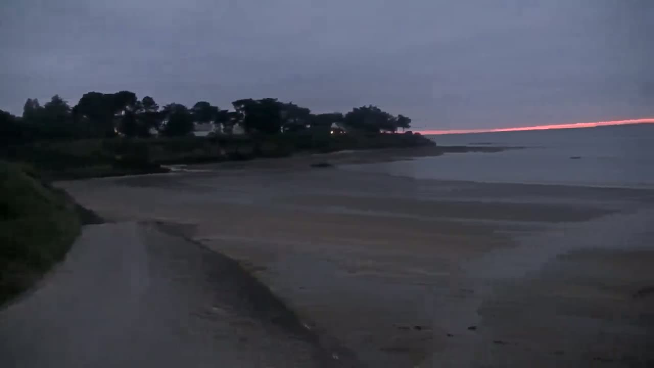 A low-light, overcast sunrise illuminates a calm bay, where a sandy beach stretches out to a treeline on a small headland.
