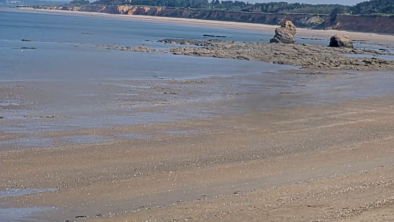 A sandy beach stretches to a calm sea, where rocky outcrops are visible at low tide, under a clear sky.