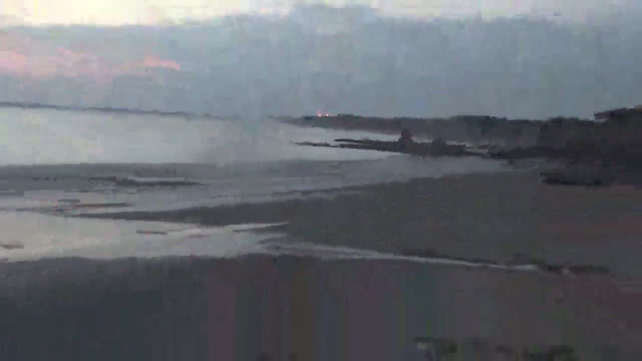 A hazy, overcast sky hangs over a calm body of water meeting a dark, sandy shoreline.