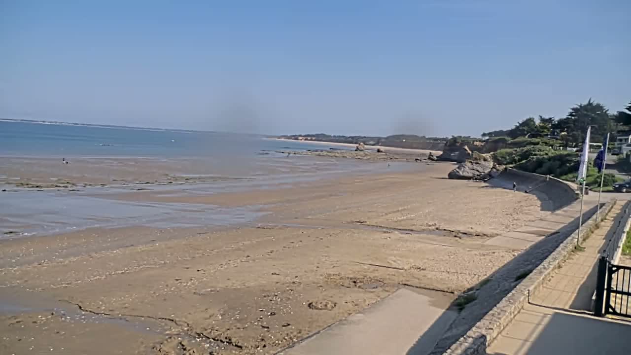 A sandy beach stretches to a calm ocean under a clear blue sky, with a low stone wall and vegetation visible along the shoreline.