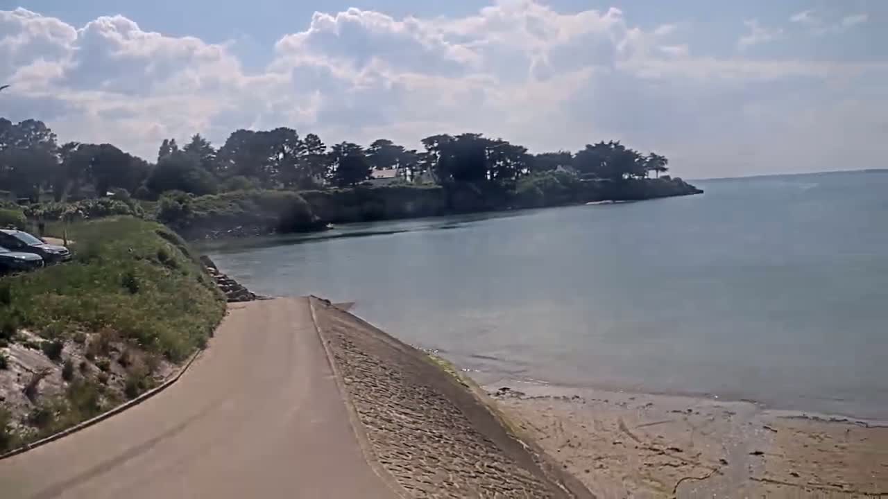 A paved road leads down to a calm, light-blue body of water with a sandy beach, under a partly cloudy sky.