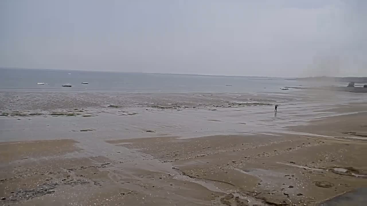 A hazy, overcast day reveals a wide sandy beach with shallow water, a lone person walking, and several small boats in the distance.