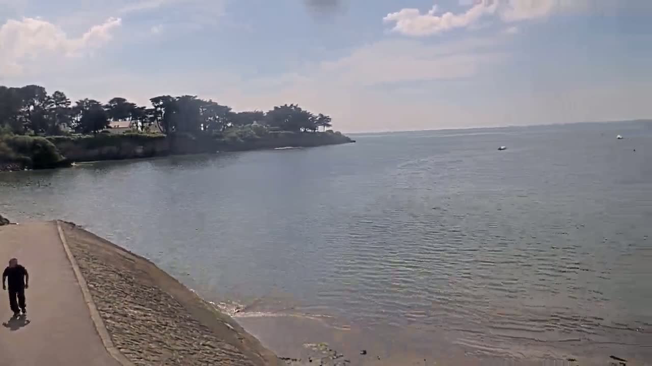A man walks on a paved path next to a calm body of water under a partly sunny sky, with trees and vegetation visible on the far shore.