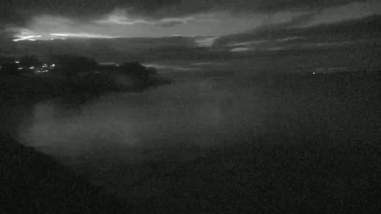 A nighttime, low-light view shows a body of water with mist or fog rising from it under a partly cloudy sky.