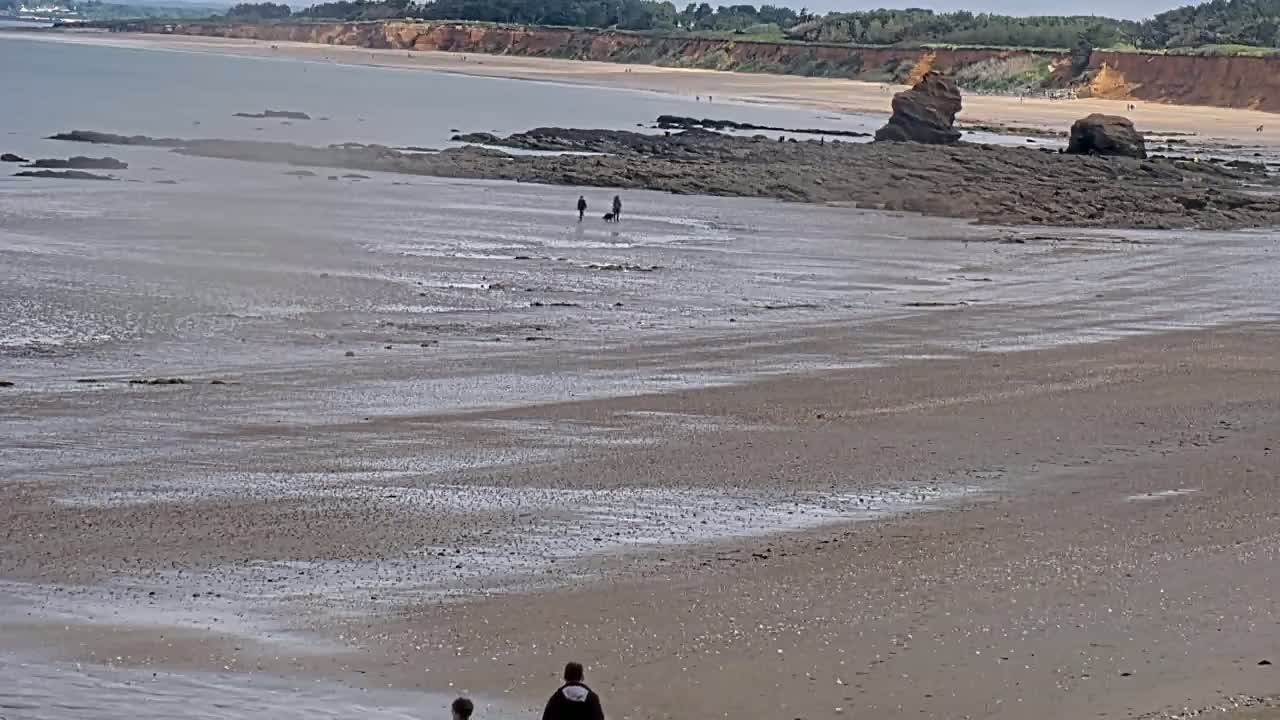 A wide sandy beach stretches to a calm sea, with a few people walking near some dark rocks and a distant cliff face.