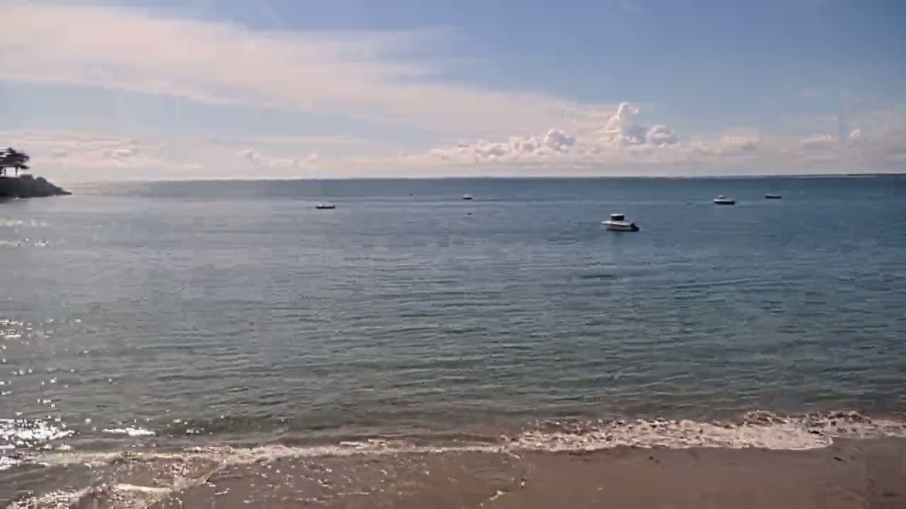 A calm, partly cloudy day shows a sandy beach with gentle waves lapping the shore, and several small boats are visible on the relatively still water of a bay.