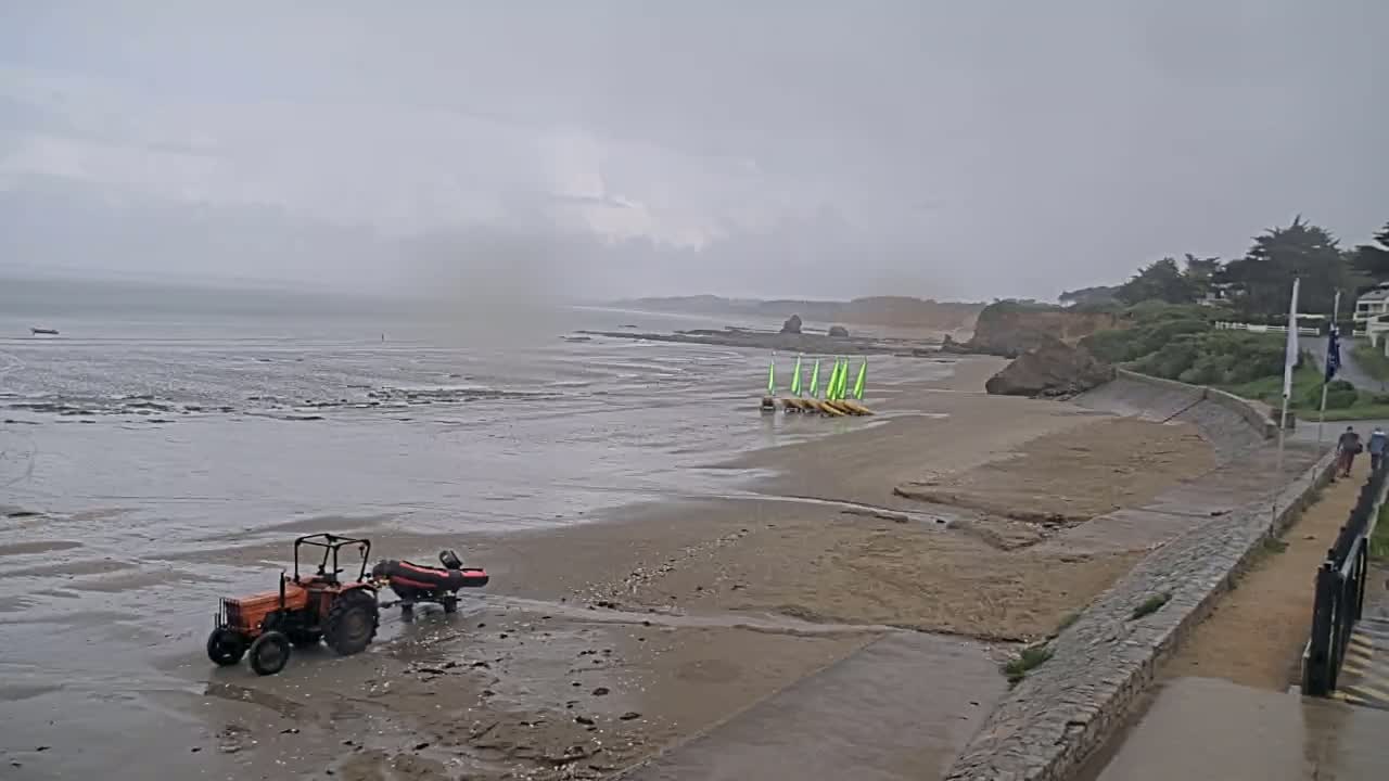 A tractor pulling a small boat sits on a wet, sandy beach under an overcast sky near a rocky shoreline and several small sailboats.