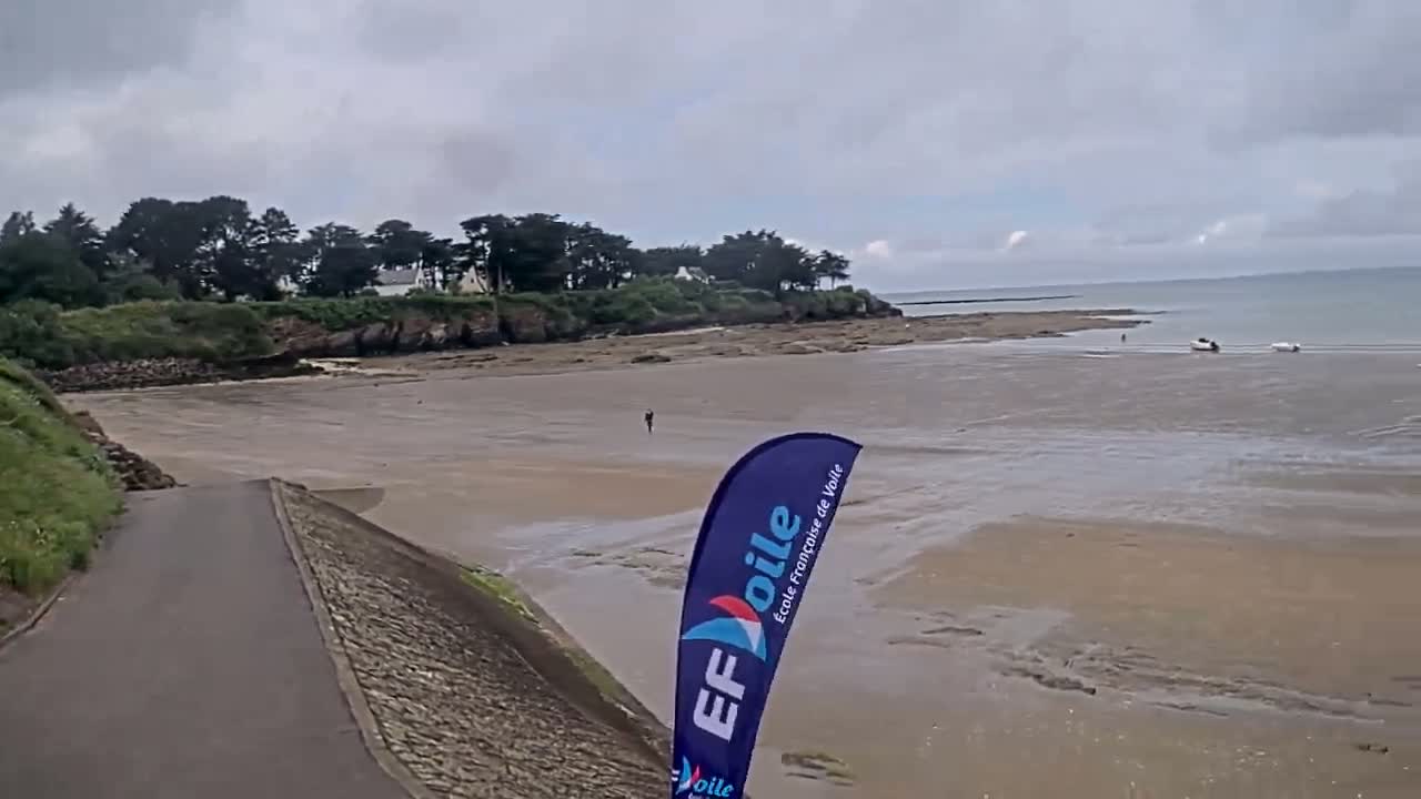 A wide sandy beach stretches out to a calm sea under an overcast sky, with a small group of trees and houses visible in the background.