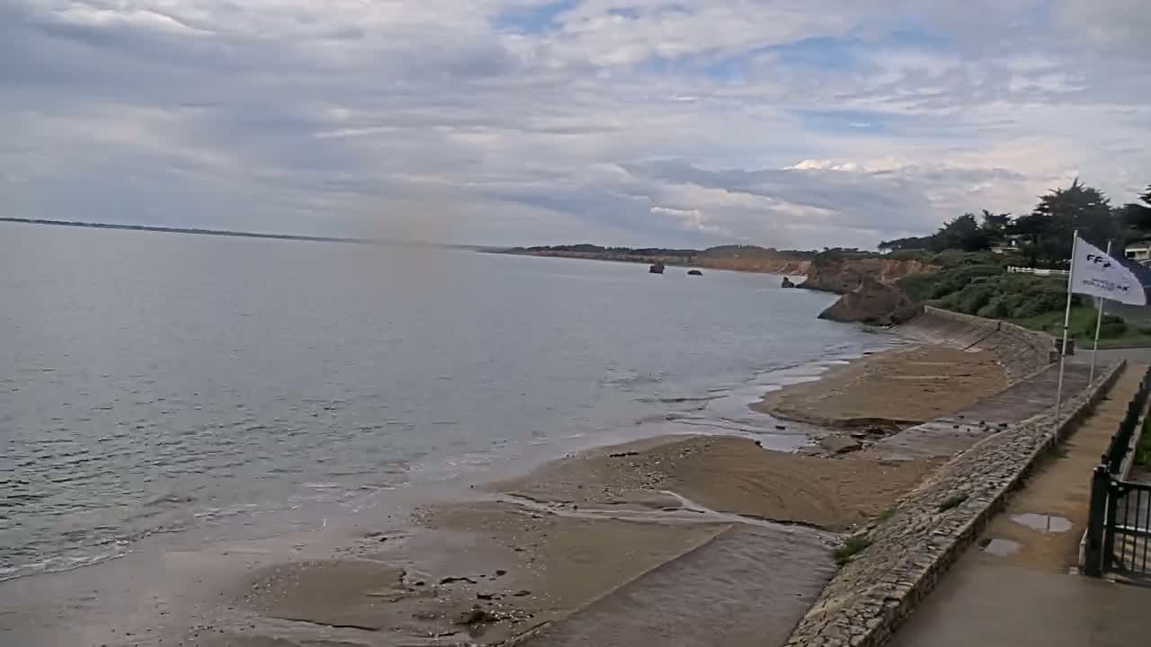 A sandy beach stretches along a calm, grey sea under a cloudy sky, backed by a low cliff with some vegetation and a paved walkway.