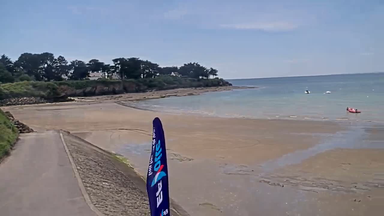A sandy beach with calm, light-blue water stretches out from a paved road bordered by green vegetation and a line of trees under a mostly sunny sky.