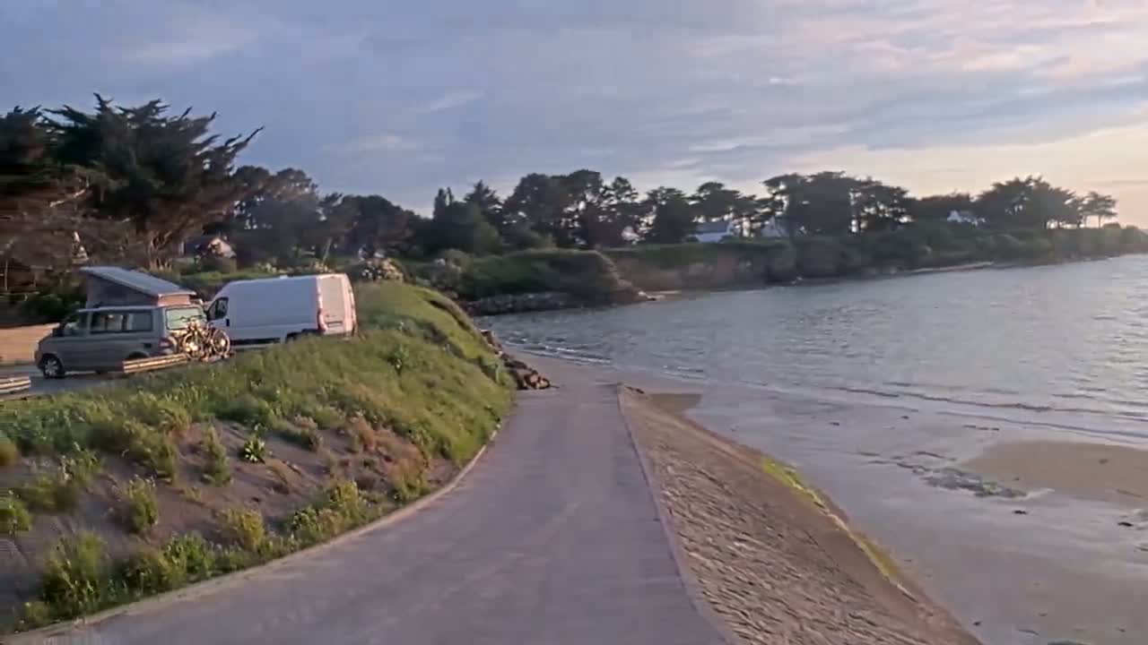 A paved road leads down to a calm, sandy beach and bay, with two vans parked nearby under a partly cloudy sky.