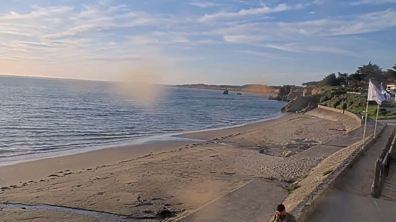 A sandy beach with gentle waves under a partly cloudy sky stretches alongside a paved walkway, backed by a low cliff with sparse vegetation.