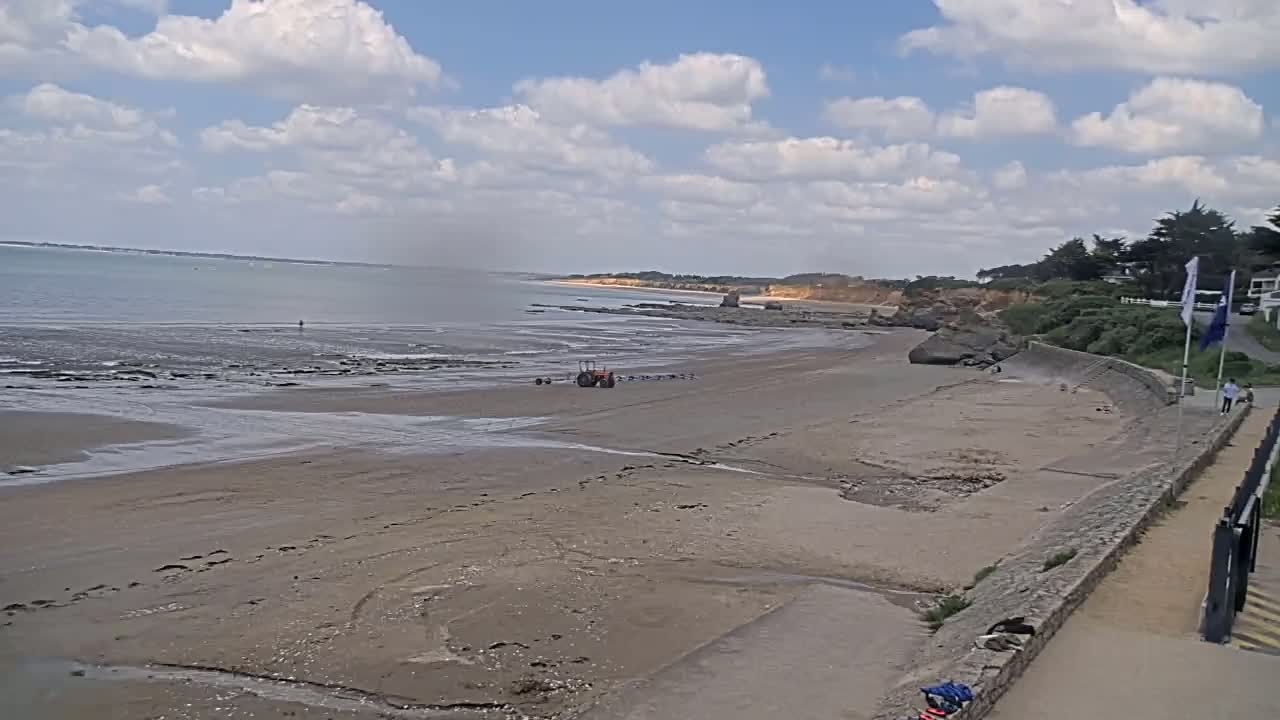 A tractor sits on a sandy beach next to a calm sea under a partly cloudy sky.