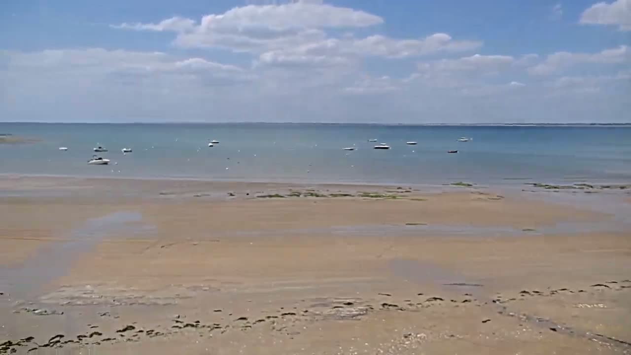 A sandy beach stretches towards a calm, light-blue body of water under a mostly sunny sky with scattered clouds, where several small boats are anchored.