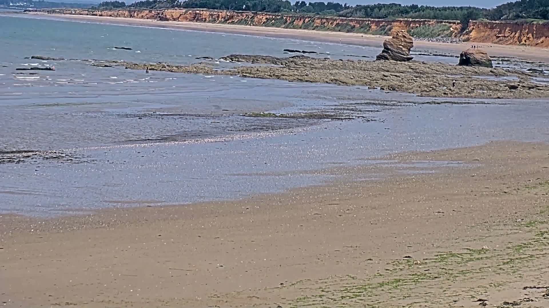 A sandy beach with calm, shallow water reveals exposed rocks and a distant, red-cliffed coastline under a sunny sky.