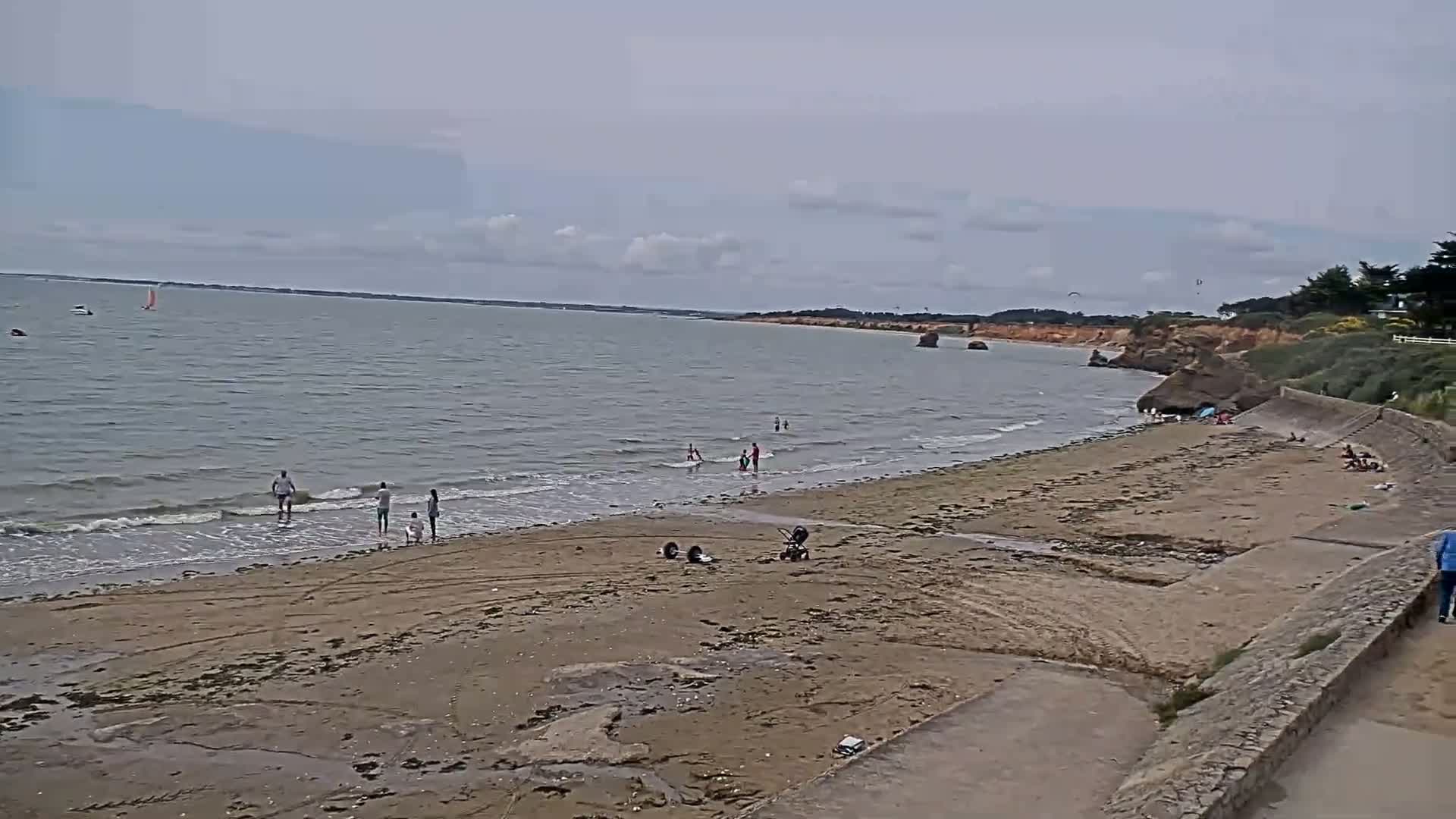 A sandy beach with a few people on it stretches along a calm, gray sea under a mostly cloudy sky.