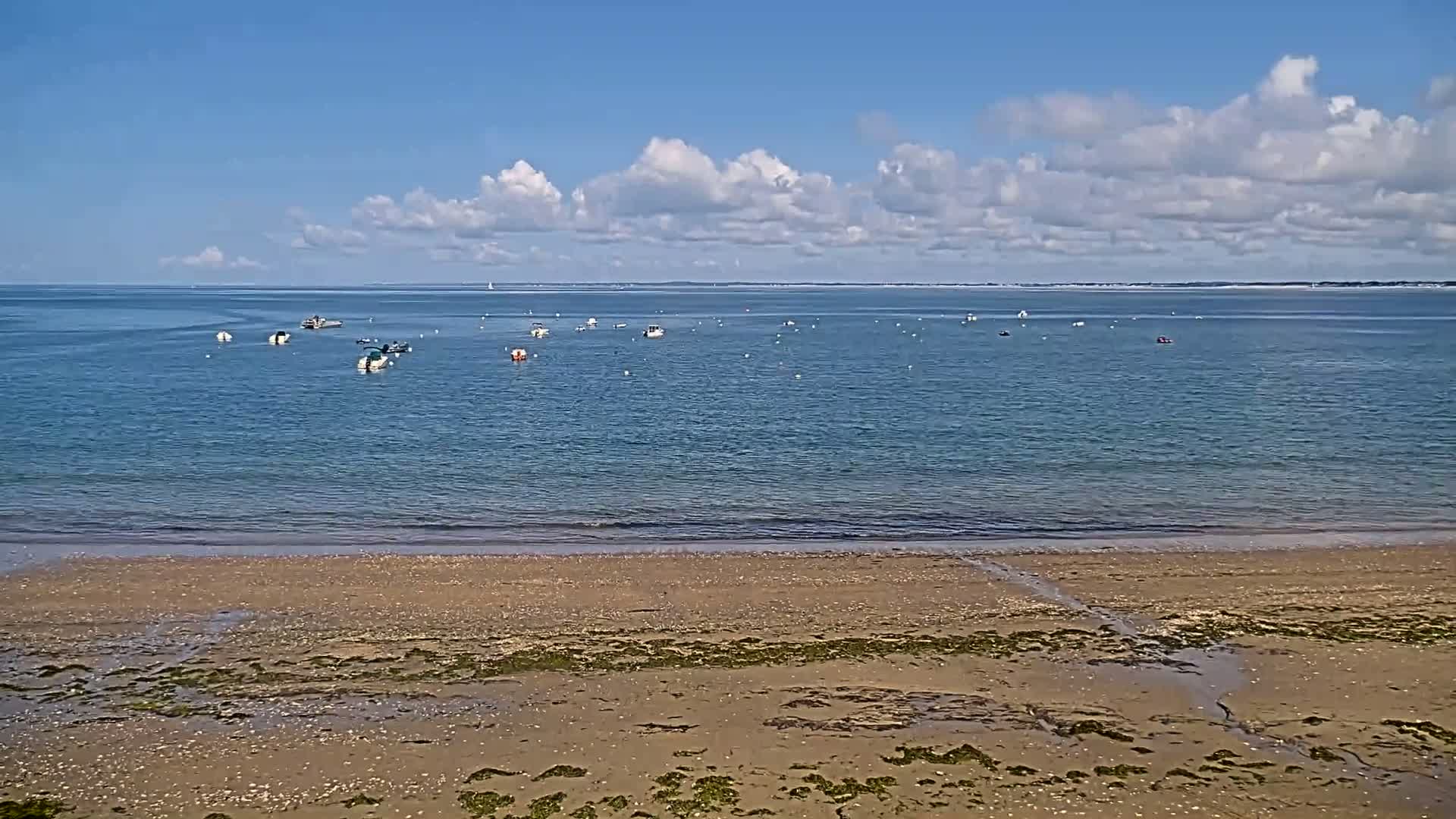 A sandy beach stretches to a calm, blue ocean dotted with small boats under a mostly sunny sky with scattered clouds.