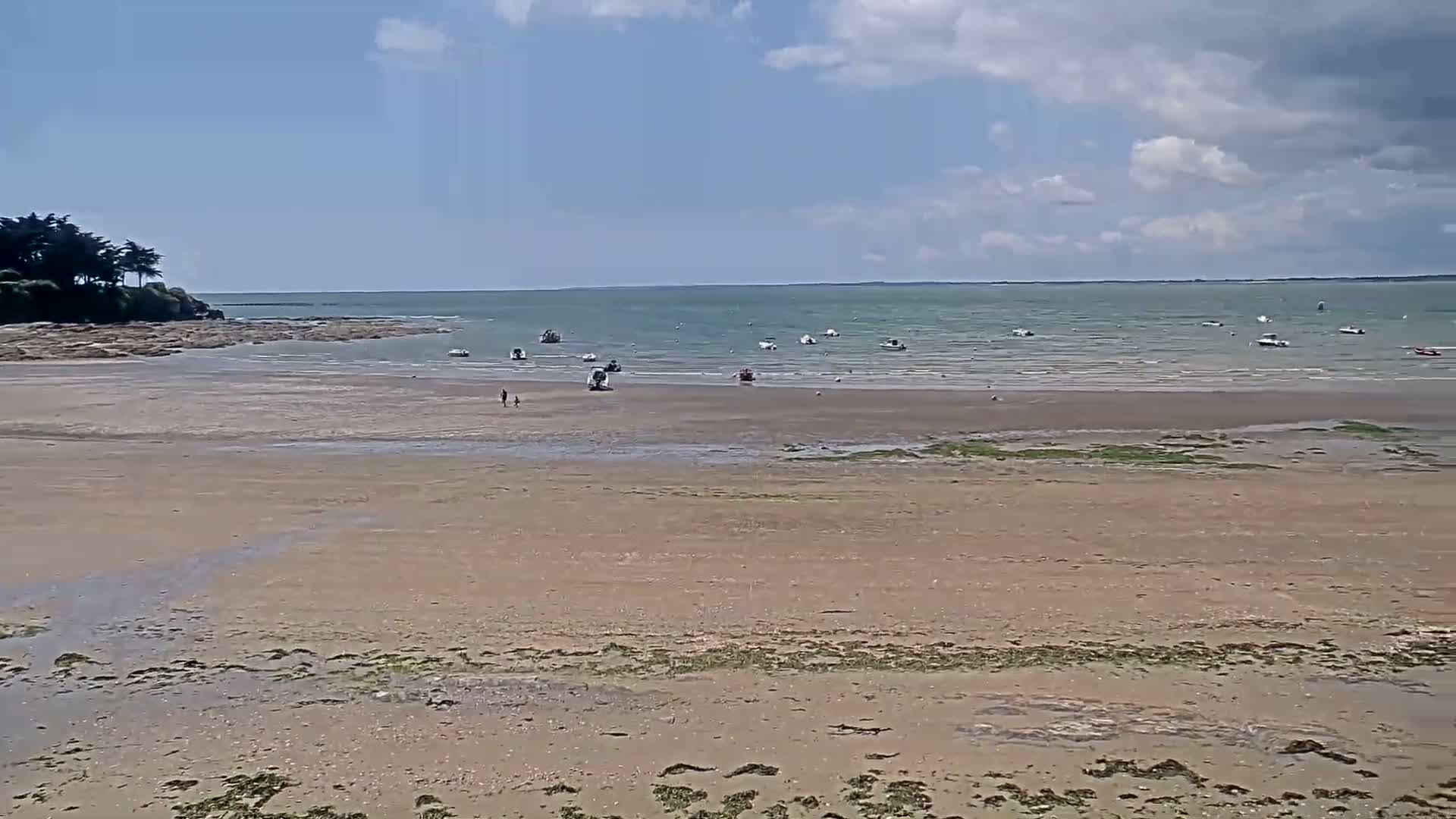 A wide sandy beach stretches to a calm sea dotted with small boats under a partly cloudy sky.