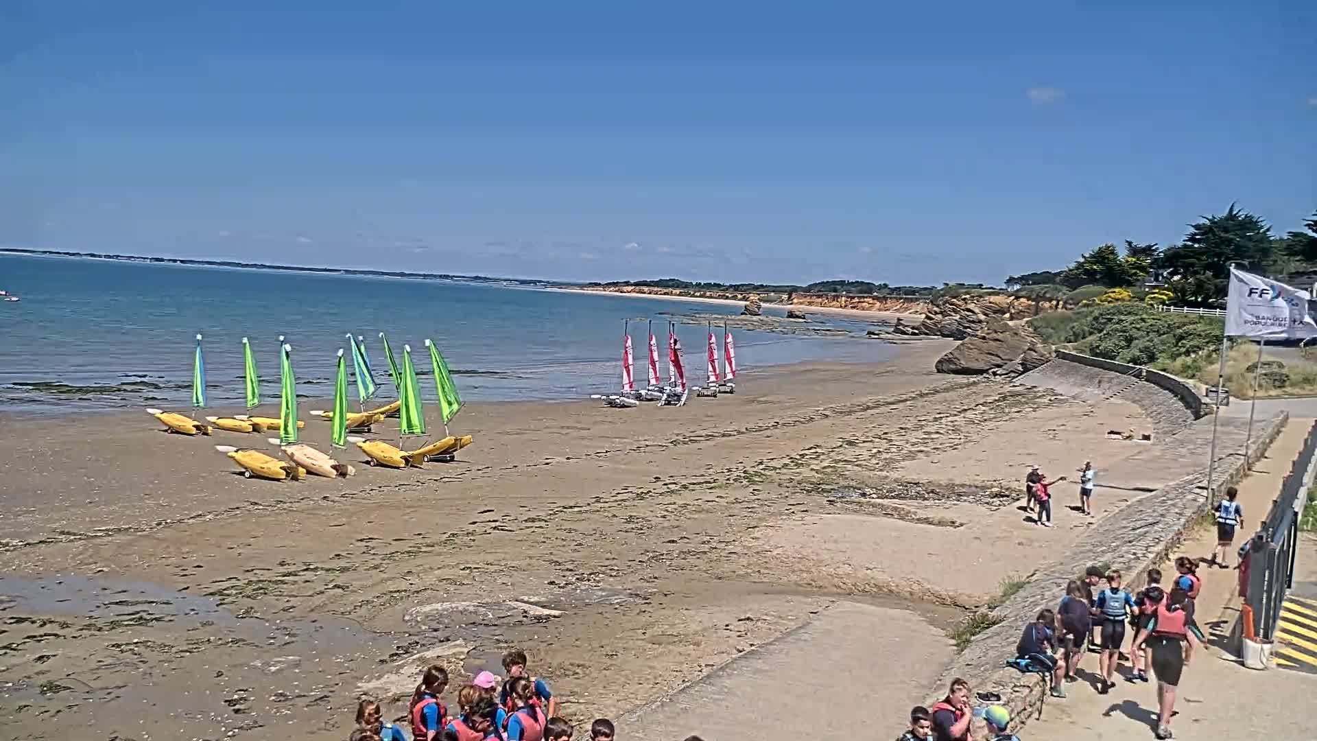 A sandy beach with numerous small sailboats and groups of children, under a clear blue sky.