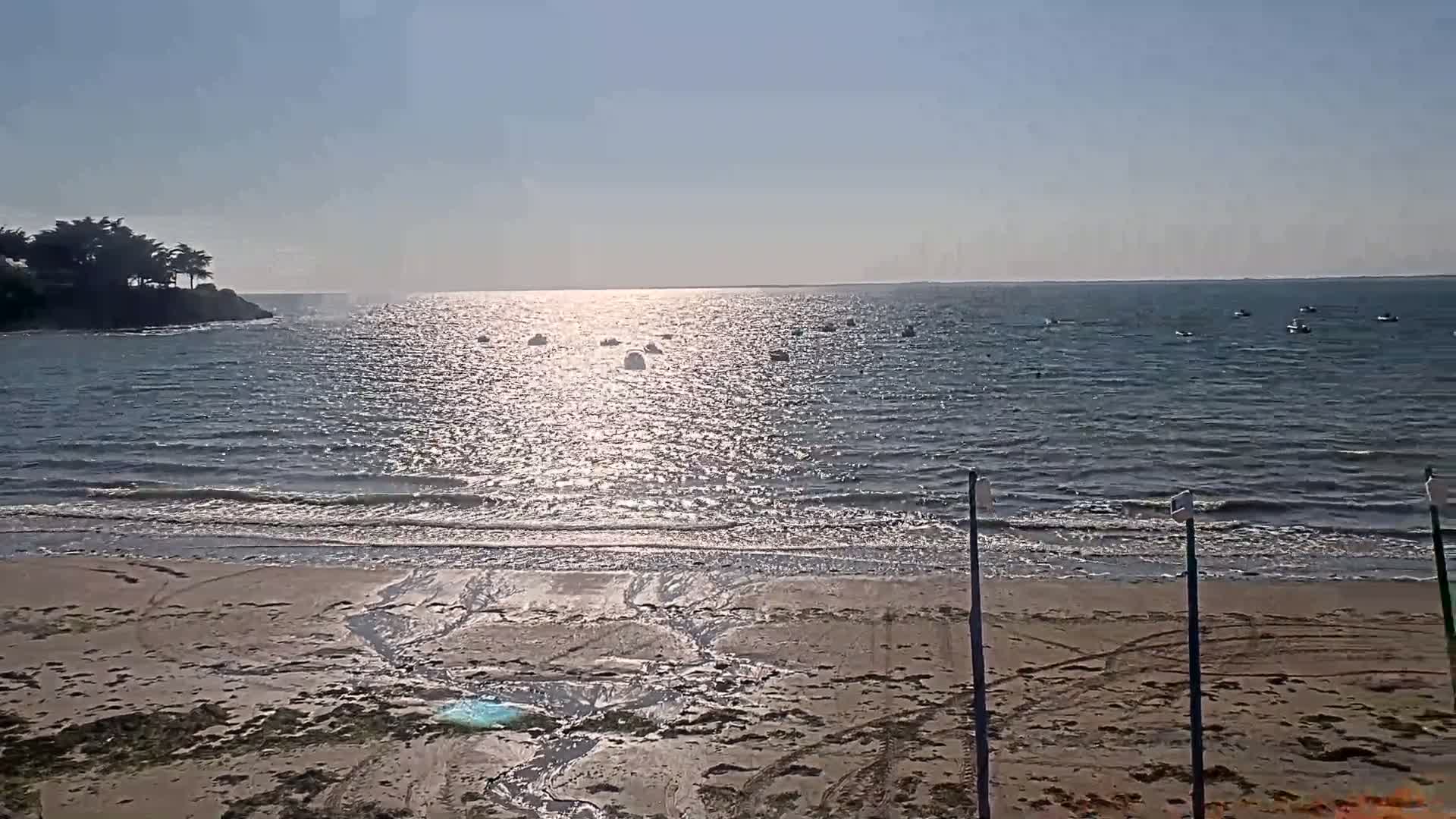 A sandy beach with gentle waves laps the shore under a bright, sunny sky, with several small boats visible on the calm water.