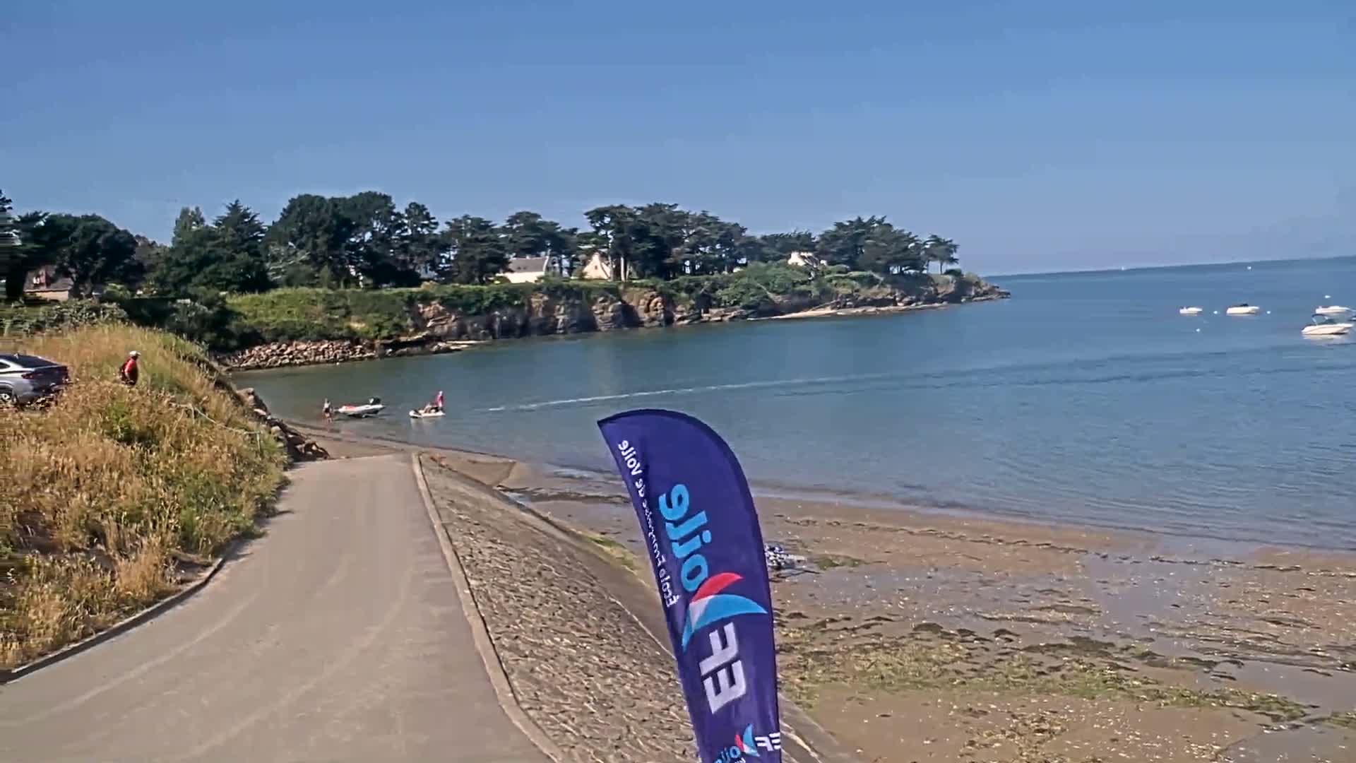 A paved boat ramp leads to a calm bay under a clear blue sky, where several small boats are moored and people are launching watercraft.