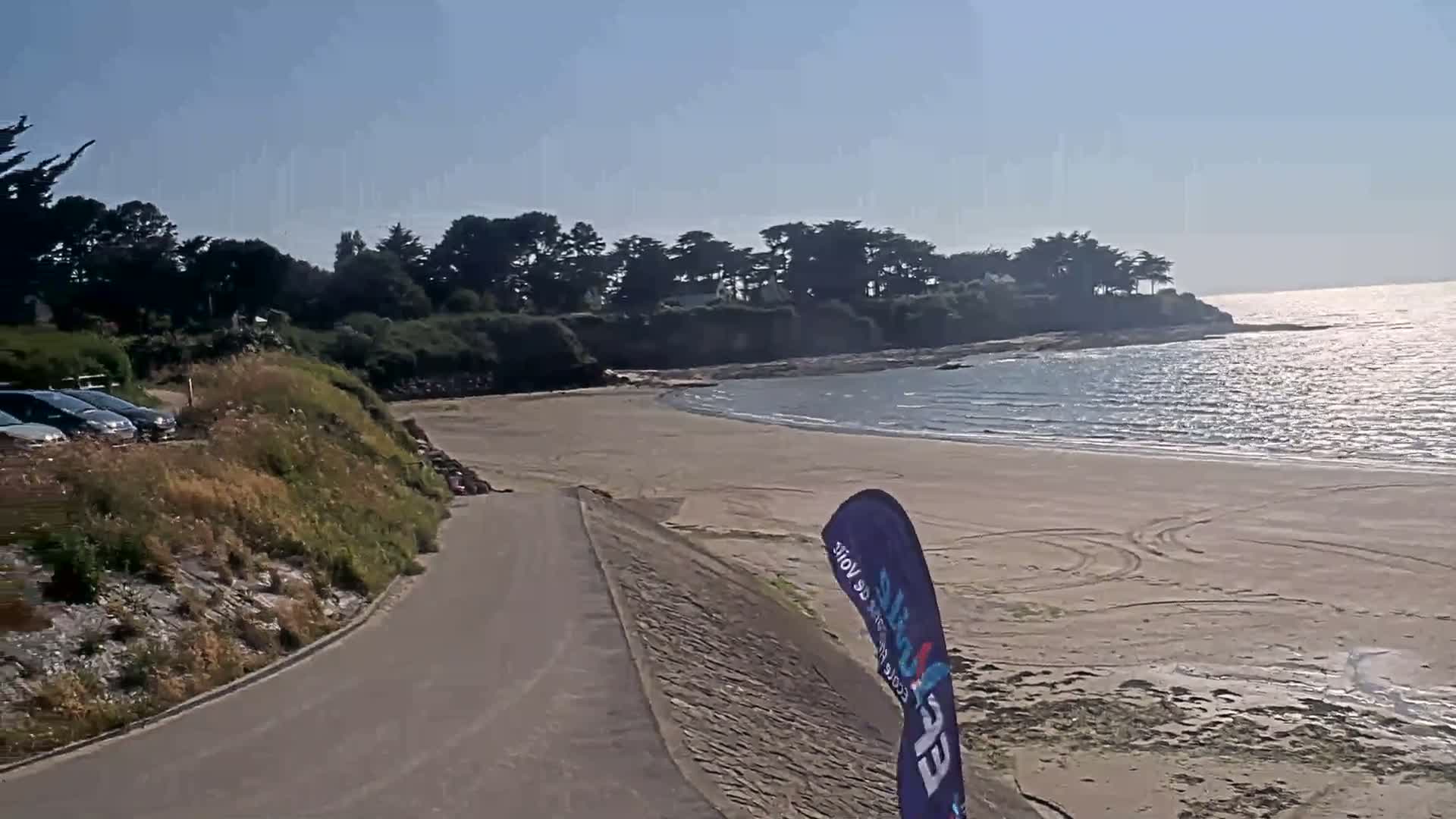 A paved path leads to a sandy beach curving around a calm, sunlit ocean bordered by green vegetation and a few parked cars.