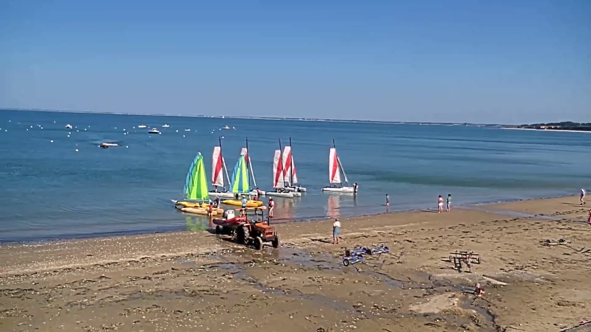 A sandy beach on a sunny day with several small sailboats in the calm water, a tractor on the shore, and a few people on the beach and in the water.