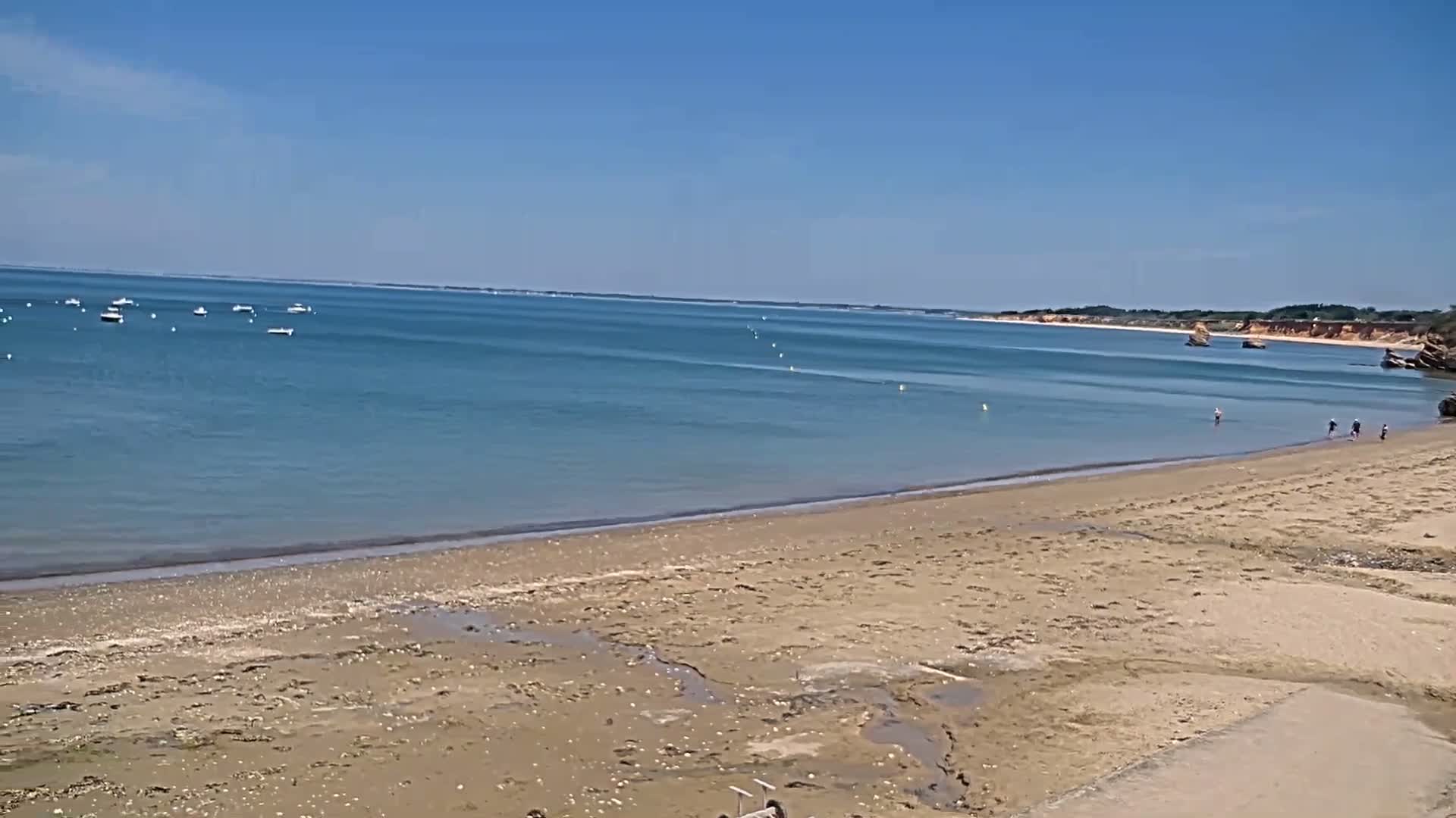 A sandy beach stretches to a calm, blue ocean under a clear, sunny sky, with several small boats visible in the distance and a few people near the water's edge.