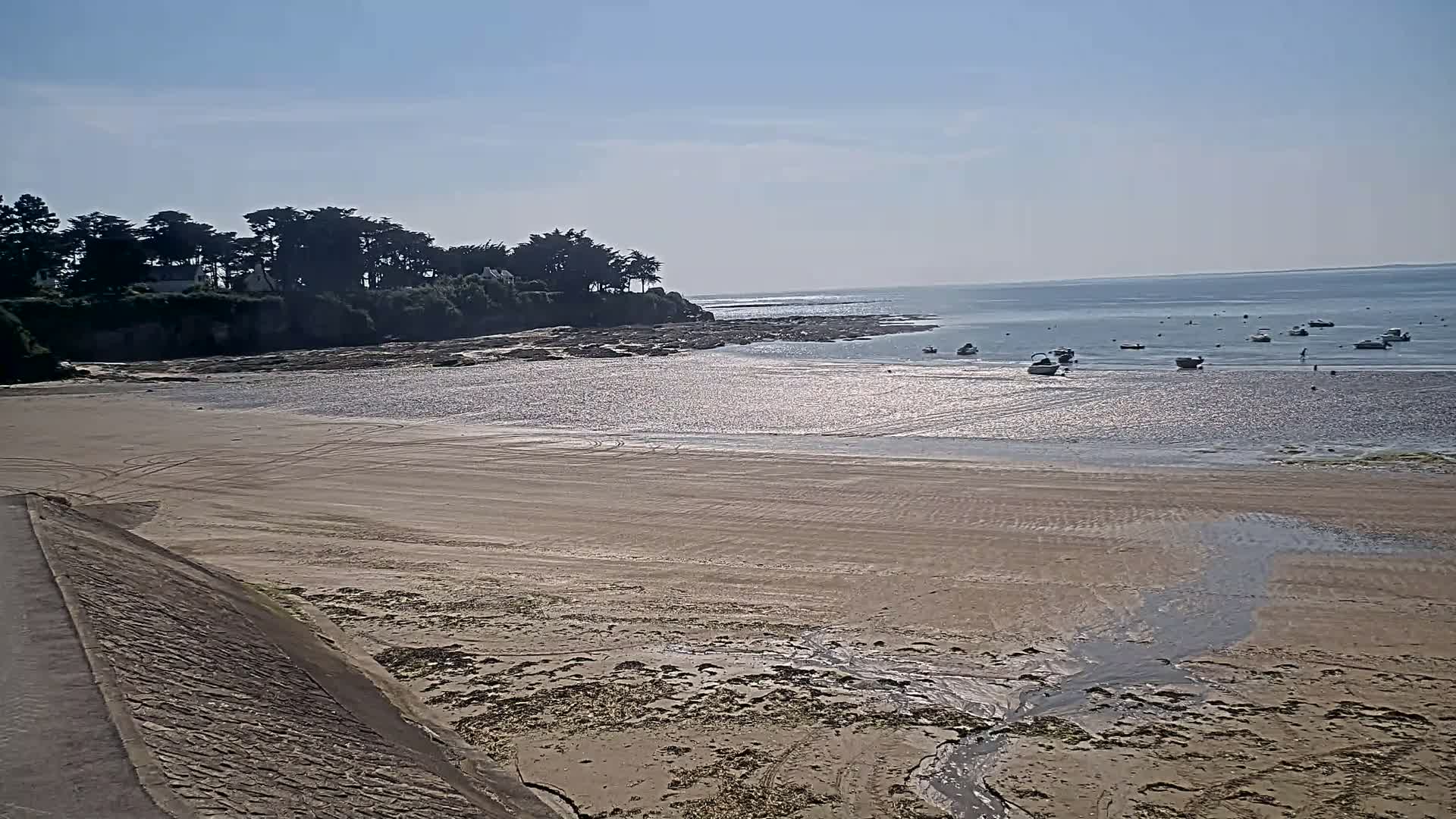 A sandy beach with tire tracks stretches towards a calm body of water where several small boats are anchored, all under a clear, sunny sky.