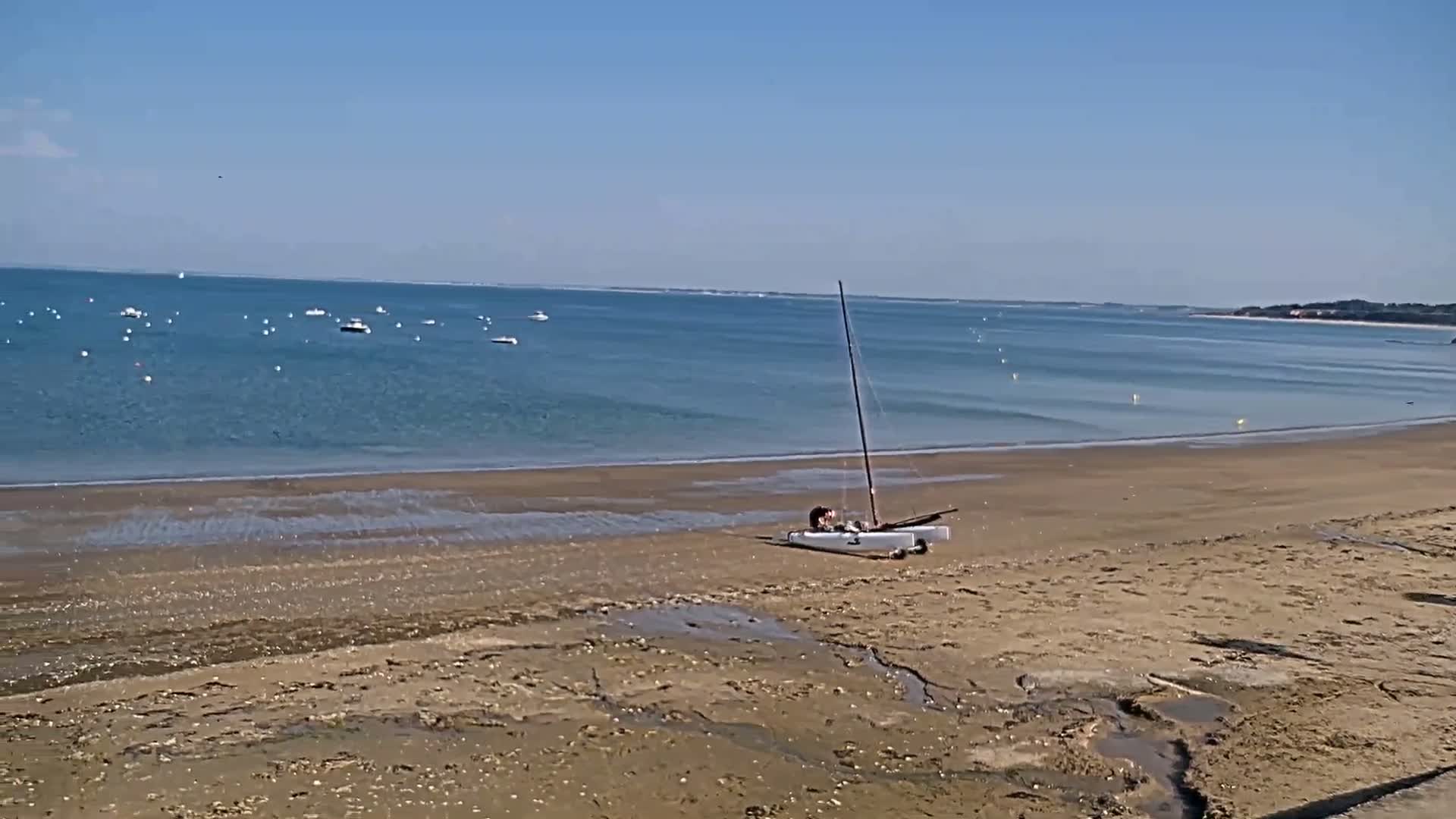 A small sailboat sits on a sandy beach next to calm, blue water dotted with small boats under a clear blue sky.