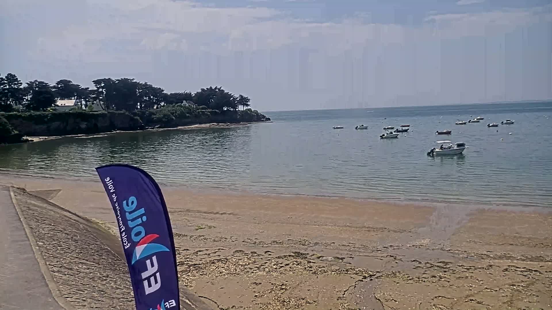 A sandy beach and calm, shallow water with several small boats are visible on a partly cloudy day, backed by a line of trees and houses on a small cliff.
