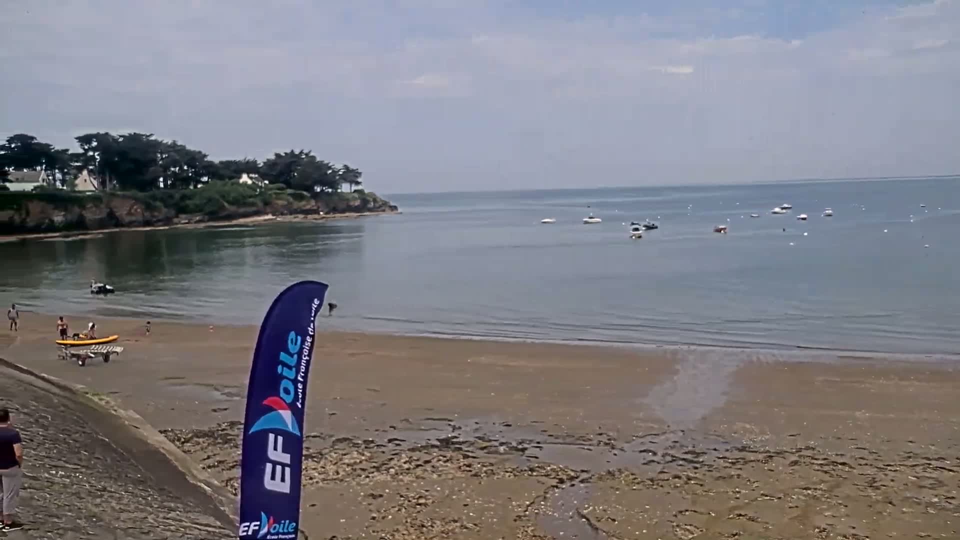 A sandy beach with several people and a few small boats on calm water under a mostly clear sky.