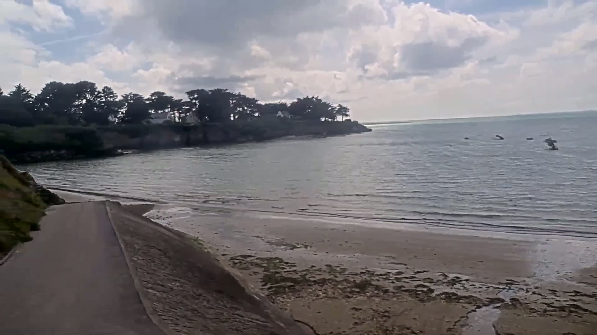 A partly cloudy day reveals a calm bay with a sandy beach and a paved path leading down to the water, bordered by trees and vegetation.