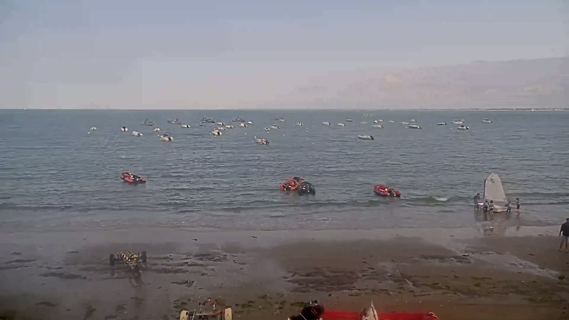 Numerous small boats are scattered across a calm body of water near a sandy beach where several people are near a small sailboat.