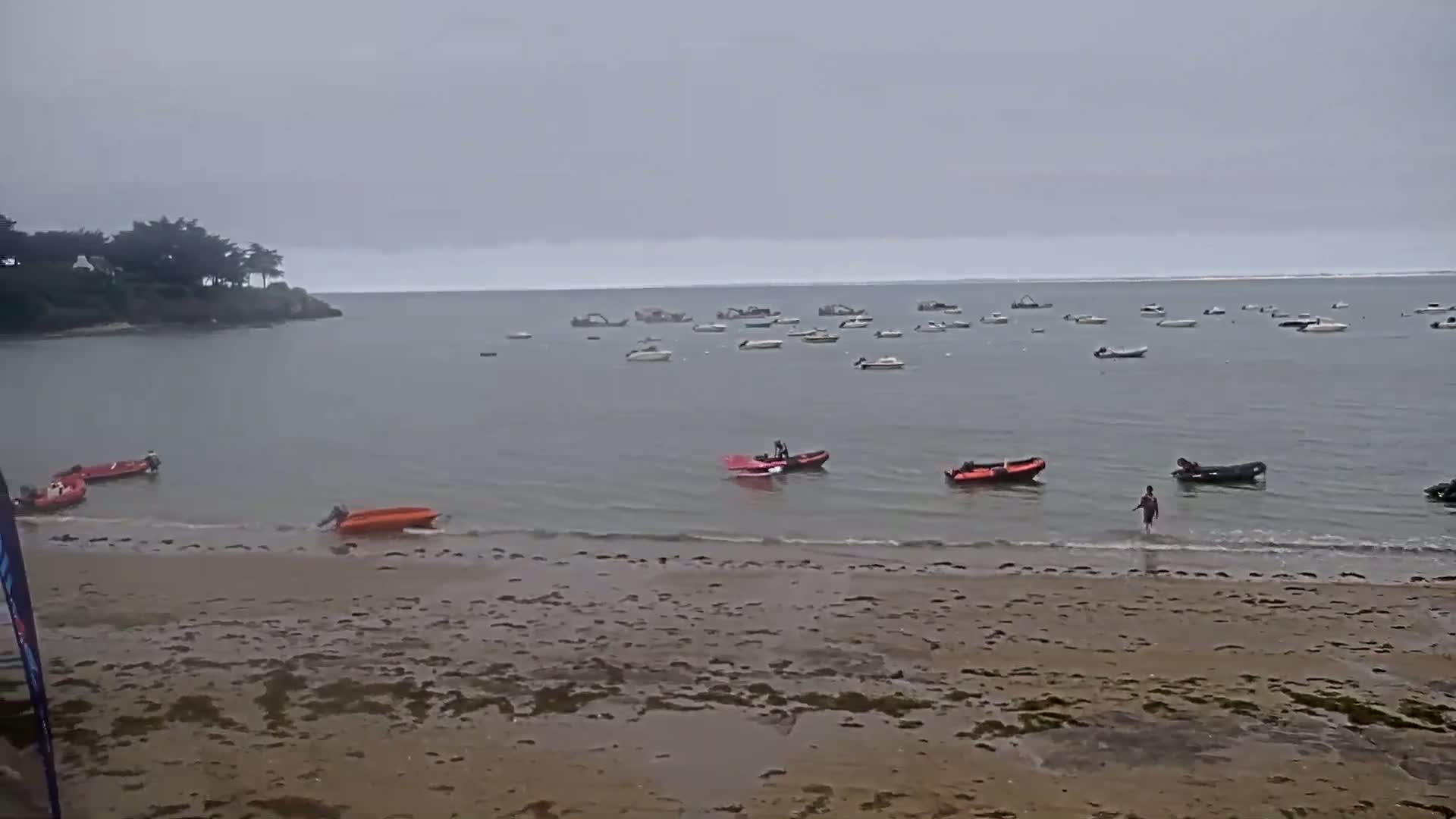 A sandy beach with several small boats in the calm, overcast water, and a person wading into the sea near the shore.