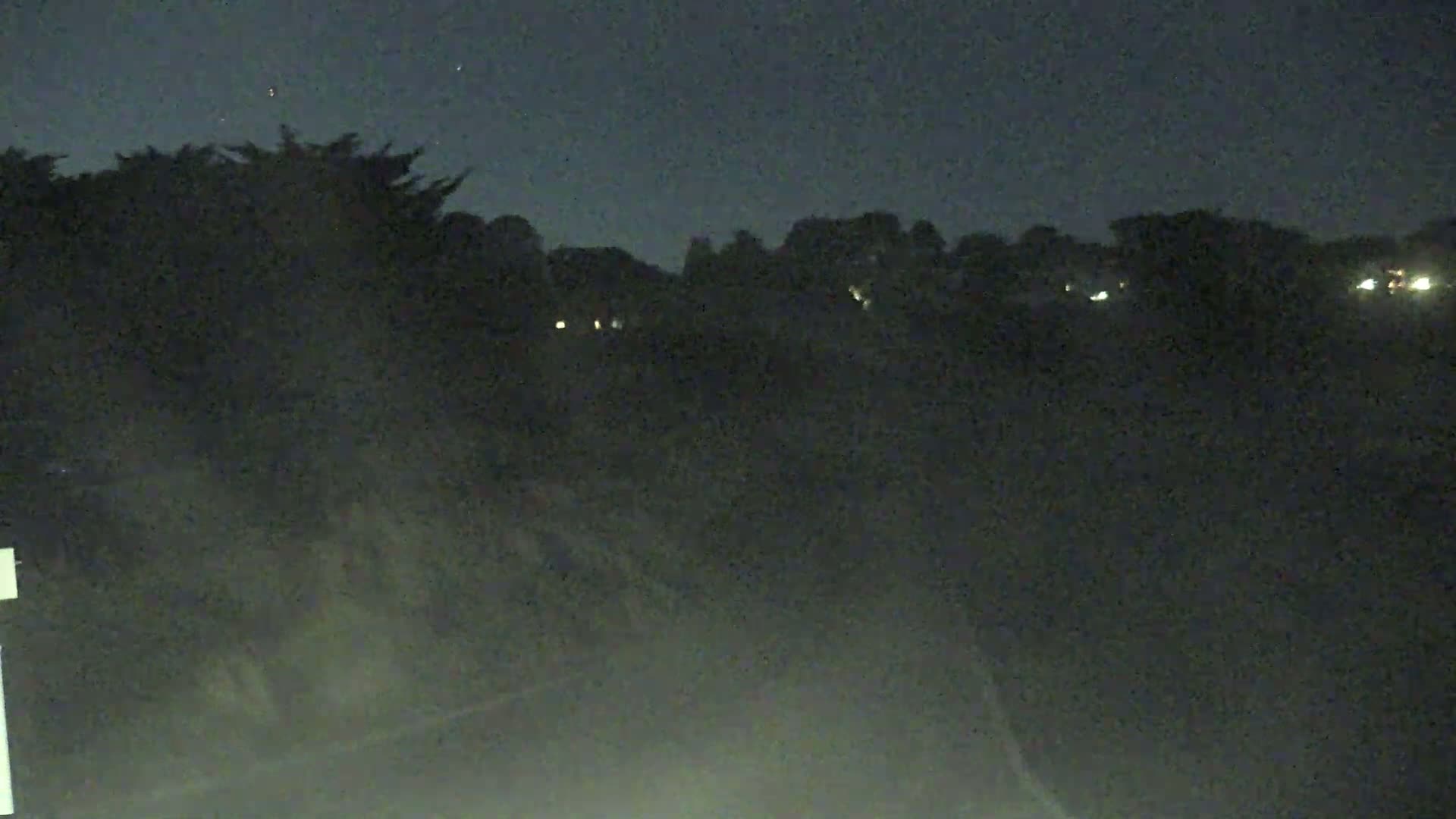 A nighttime view shows dark trees and a distant line of houses with faint lights under a dark, clear sky.