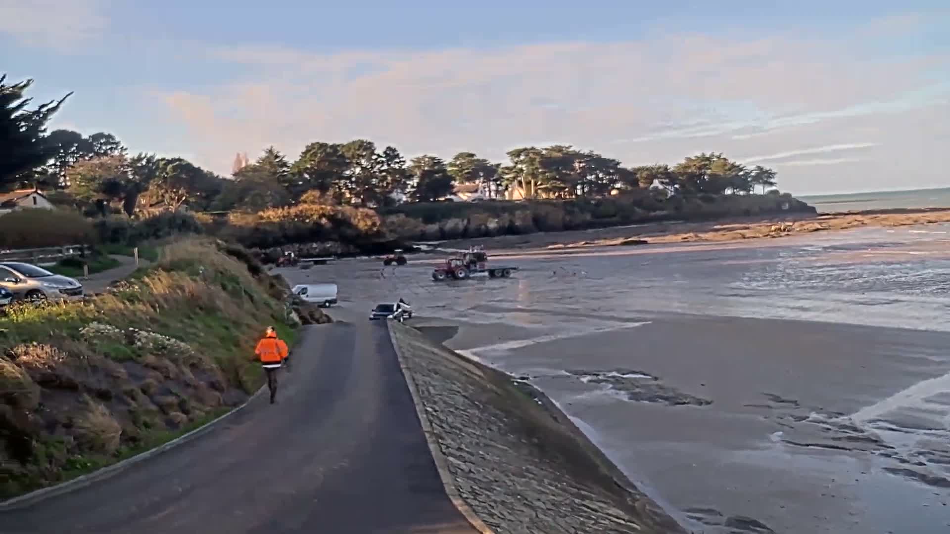 A person in an orange jacket walks down a paved road next to a grassy hill overlooking a muddy, exposed beach with tractors and vehicles, while a coastal village and the ocean lie under a partly cloudy sky.
