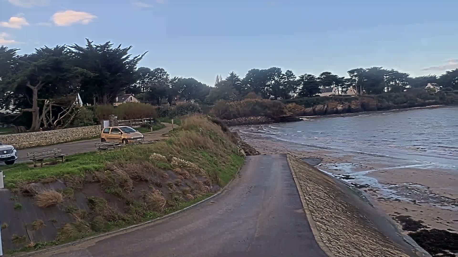 A coastal road with parked cars and benches runs alongside a grassy slope leading to a sandy bay with gentle waves, all under a partly cloudy sky, with houses and dense trees visible along the distant shoreline.
