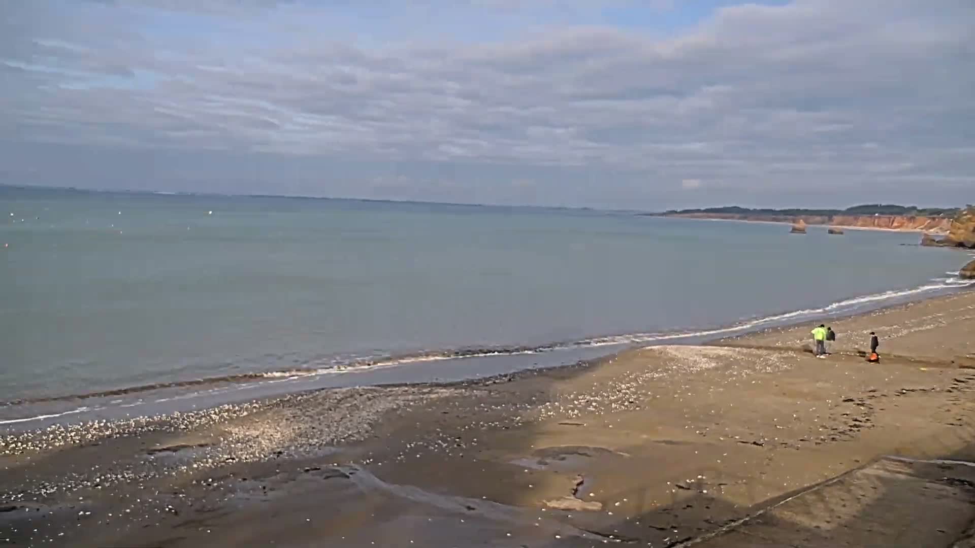 A coastal road with parked cars and benches runs alongside a grassy slope leading to a sandy bay with gentle waves, all under a partly cloudy sky, with houses and dense trees visible along the distant shoreline.